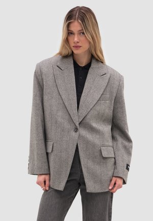 Lyshåret kvinde iført en oversized grå blazer over en sort skjorte med krave og mørkegrå jeans, stående mod en ensartet lys baggrund.