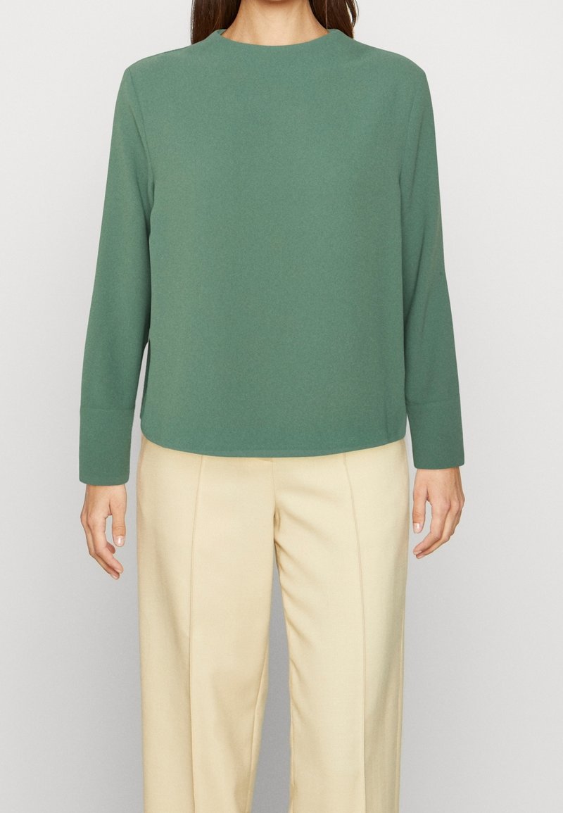 Femme portant un haut vert sauge à manches longues et un pantalon beige taille haute, debout devant un fond clair uni.