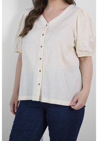 Blouse beige clair avec des manches courtes bouffantes, fermeture à bouton sur le devant et décolleté en V. Fabriquée dans un tissu texturé, elle présente un ourlet droit.