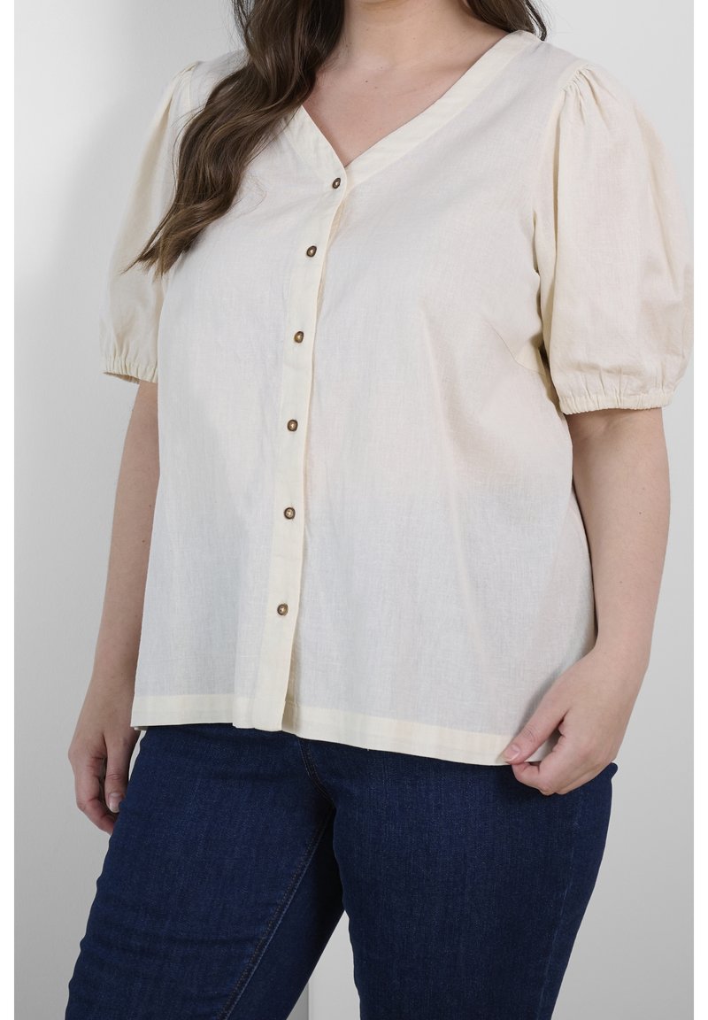 Blouse beige clair avec des manches courtes bouffantes, fermeture à bouton sur le devant et décolleté en V. Fabriquée dans un tissu texturé, elle présente un ourlet droit.