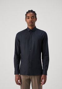 Schwarz langärmliges Shirt aus glattem Stoff mit einem Button-Down-Kragen, Frontknöpfen und taillierter Passform. Getragen mit hellen Hosen.