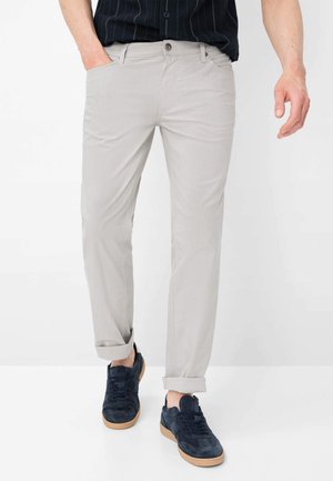 Mand iført lyse beige bukser, der er rullet op, sort skjorte med nålestriber og mørkeblå sneakers med gummisåler, stående mod en hvid baggrund.