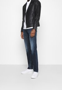 Giacca da motociclista in pelle nera sopra una t-shirt bianca, abbinata a jeans blu e scarpe da ginnastica bianche. Caratteristiche includono cerniere e un taglio classico.