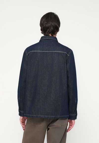Veste en denim bleu foncé avec un col, des manches longues et des coutures visibles. Présente une coupe droite et des détails de boutons aux poignets.