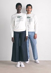 Due modelli indossano felpe bianche Lacoste e sneakers bianche, uno con jeans scuri larghi e l'altro con jeans azzurri chiari, si trovano di fronte a una parete bianca.