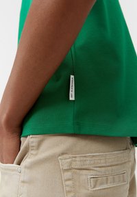 Persona vestida con una camiseta verde que tiene una etiqueta que dice "EST. EN ESTOCOLMO" y pantalones beige, con la mano descansando en el bolsillo delantero.