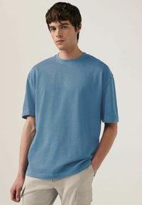 Tricou supradimensionat bleu deschis, cu guler rotund, mâneci scurte și textură moale. Puratat cu pantaloni cargo de culoare deschisă.