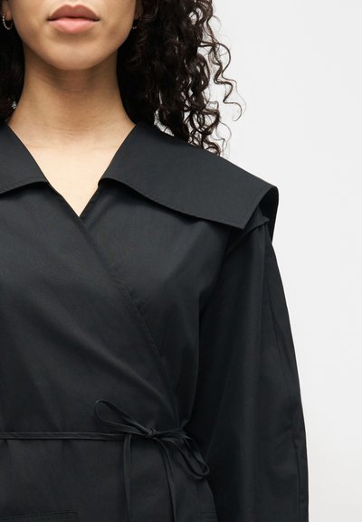 Γυναίκα που φορά μαύρο φόρεμα τύπου wrap με φαρδύ γιακά, μακριά μανίκια και λεπτή ζώνη στη μέση, πάνω σε απλό φόντο.