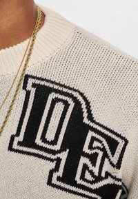 Strickpullover in Creme mit großem schwarzem "DE"-Logo, ausgestattet mit einem gerippten Rundhalsausschnitt und einer glatten Textur. Goldkette im Bild sichtbar.