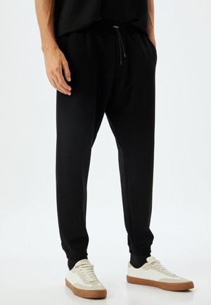 Sorte joggebukser med elastisk talje og ankelflader, med sidelommer og afslappet pasform, parret med hvide sneakers.