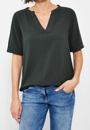 Femme portant un chemisier à manches courtes texturé vert foncé avec un col en V et un jean bleu, une main dans la poche.