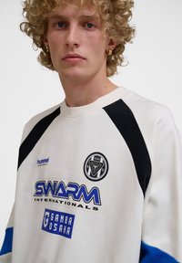 Vitt sweatshirts med svarta och blå accenter, med en rund logotyp och tryckt text. Tillverkad av ett mjukt, strukturerat material.