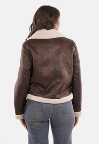 Chaqueta de cuero marrón con cuello y puños de borreguito, con un diseño ajustado y detalles de costura vertical, llevada sobre jeans grises.