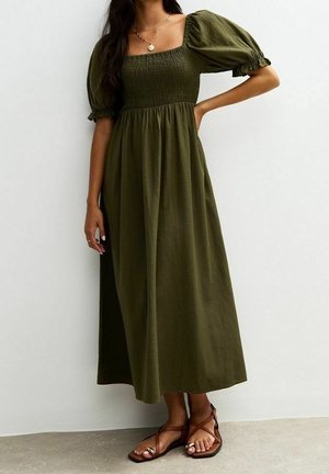 Femme portant une robe vert olive à manches bouffantes, longueur cheville, avec un corsage smocké et des sandales plates marron à brides.