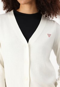 Cardigan couleur crème avec un col en V et cinq boutons. Présente un petit logo triangulaire rouge sur la poitrine et des poignets côtelés. Texture lisse.