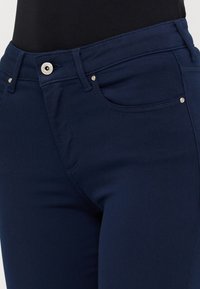 Marineblå jeans laget av elastisk stoff, med glidelås og knappelukking, samt frontlommer og metalldetaljer.