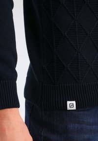 Ciemnoniebieski dziergany sweter z diamentowym wzorem, prążkowanym dołem i białą metką marki, na ciemnoniebieskich dżinsach, noszony przez osobę.