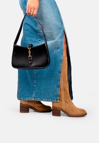 Bottes en suède beige hauteur genou avec des talons carrés, associées à une jupe en denim bleue avec une fente sur le côté et un sac à main noir avec des détails en or.