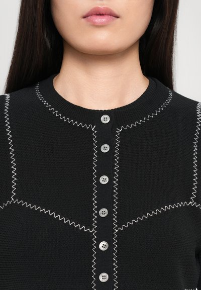 Cardigan negru din tricot cu guler rotund, prezentând cusături în zigzag albe contrastante și cinci nasturi argintii pe partea din față.