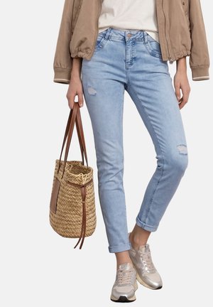 Person trägt hellblaue zerrissene Jeans, beige Bomberjacke, weißes Top, beige Sneaker und hält eine geflochtene Strohtasche mit braunen Lederriemen.