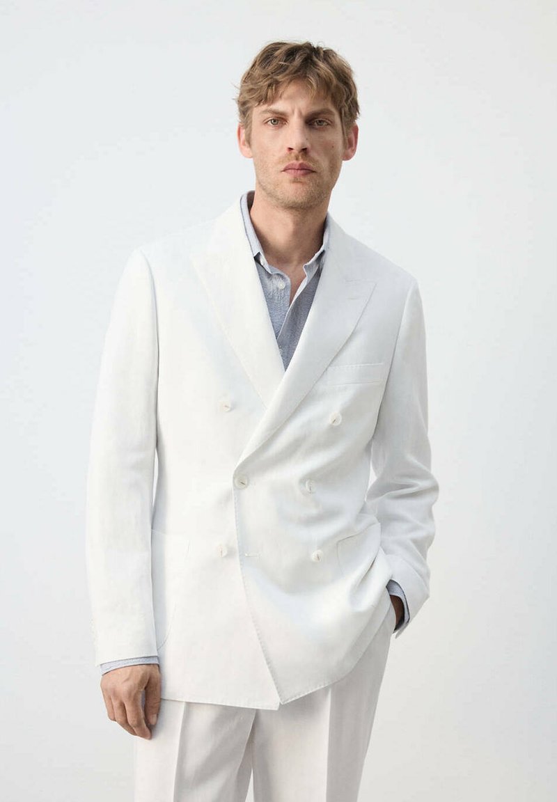 Homme aux cheveux châtains clair portant une veste de costume croisée blanche et un pantalon blanc, debout devant un fond clair uni.