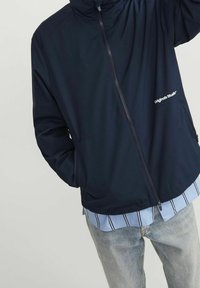 Giacca blu navy con zip realizzata in tessuto leggero, con cappuccio, tasche laterali e dettaglio logo sul petto, indossata sopra una maglia a righe.