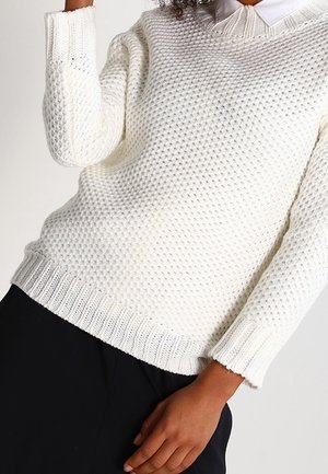 Person trägt einen strukturierten weißen Strickpullover über einem weißen Hemd mit Kragen und schwarzen Hosen, Arm erhoben, Hand locker an der Seite.