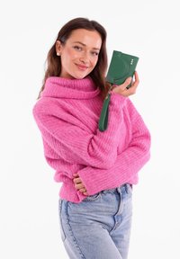 Pink knitted sweater with a high neck; model holds a small, textured green wallet with a wrist strap and three decorative dots.