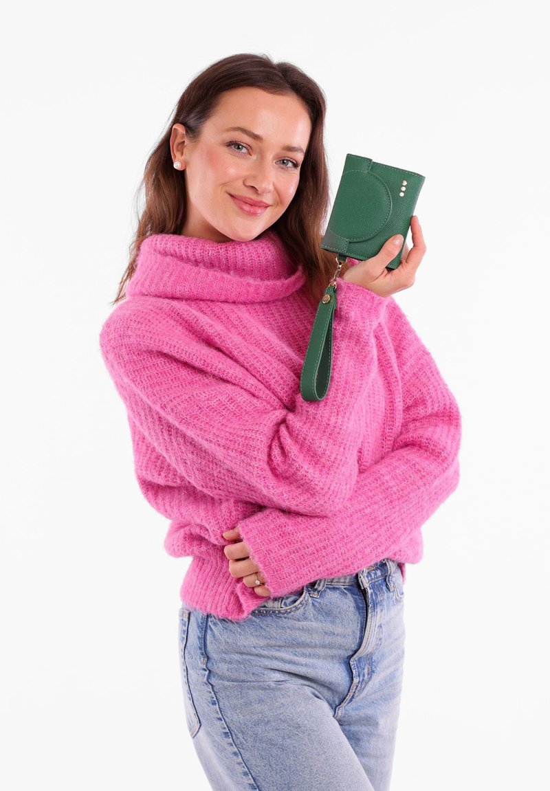 Pink knitted sweater with a high neck; model holds a small, textured green wallet with a wrist strap and three decorative dots.