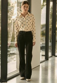 Chemise crème à boutons, ornée de pois noirs, accompagnée de pantalons noirs taille haute. L'environnement est lumineux avec de grandes fenêtres et un reflet.