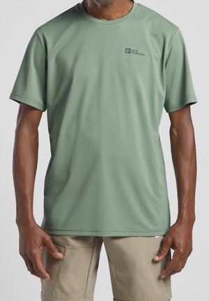 Mann trägt ein kurzärmeliges T-Shirt von Jack Wolfskin in Salbeigrün und beige Cargo-Shorts, steht mit entspannten Armen an den Seiten.