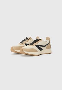Baskets beige et crème avec des accents noirs, présentant un design en daim texturé et en cuir lisse, semelles en caoutchouc avec un motif de traction en zigzag.