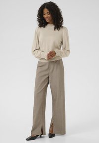 Pull en maille beige avec des manches bouffantes et des accents de boutons, associé à un pantalon gris à motifs taille haute avec des fentes latérales et des chaussures noires pointues.