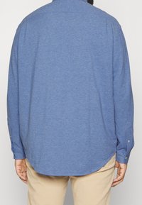 Niebieska koszula z długim rękawem i kołnierzykiem na guziki, charakteryzująca się miękką fakturą i zaokrąglonym dołem. Guziki na mankietach, bezszwowy tył.