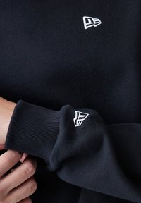 Sort sweatshirt med ribbede manchetter, prydet med et lille hvidt broderet logo på brystet og ærmet. Blødt, struktureret stof.