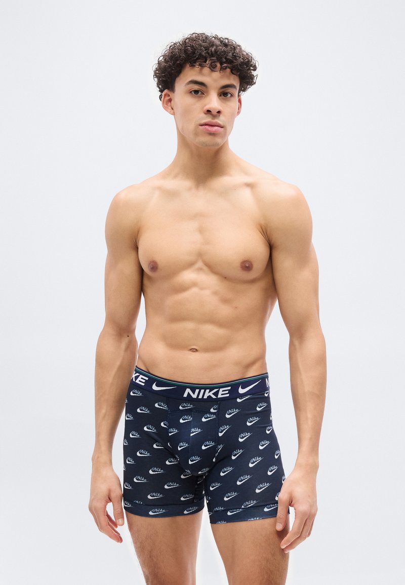 Νεαρός άνδρας με σγουρά μαλλιά, φορώντας σκούρα μπλε boxer briefs της Nike, στέκεται μπροστά σε απλό ανοιχτόγριζο φόντο.