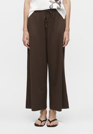 Καφέ παντελόνια με φαρδιά μπατζάκια και ζώνη με κορδόνι, συνδυασμένα με μαύρα σανδάλια τύπου φλίπ φλοπ και λευκό τοπ με μπλε και χρυστό floral σχέδιο.