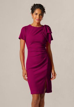 Frau mit lockigem Haar, die ein eng anliegendes, kurzärmeliges magentarotes Kleid mit einem Seitenschlitz und einem großen Schleifendetail auf einer Schulter trägt.