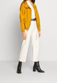 Żółta lekka kurtka z kapturem, elastycznymi mankietami, dwoma przednimi kieszeniami, zestawiona z białymi szerokimi spodniami i czarnymi botkami.