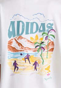 Hvit bomull T-skjorte med fargerik strandscene med fjell, palmer og figurer som spiller ball. "ADIDAS" trykt i turkis.