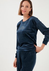 Sininen velour-kotiasu, jossa on pyöreä kaula-aukko ja pitkät hihat, sekä kultainen auringonnousukuva edessä. Pehmeä ja sileä rakenne.