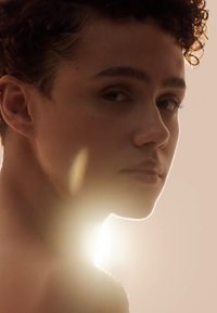 Profil av en person med kort, krøllete hår, glatt hud og en subtil øredobb, belyst av myk, diffus bakgrunnsbelysning.