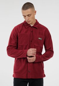 Jongeman met kort haar, gekleed in een rode fleece jas met twee borstzakken, waarvan er één een "Pro Test" logo toont, terwijl hij zijn mouw aanpast.