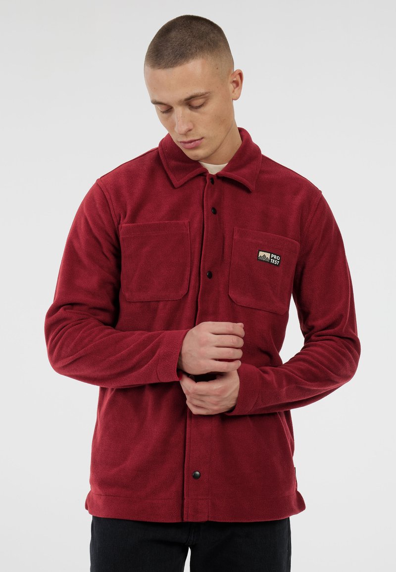 Jongeman met kort haar, gekleed in een rode fleece jas met twee borstzakken, waarvan er één een "Pro Test" logo toont, terwijl hij zijn mouw aanpast.