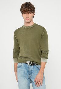 Olivzaļš džemperis ar strukturētu apdari, apaļu kakla izgriezumu un kontrastējošām gaišākām aprocēm. Valkāts ar gaiši ziliem džinsiem un sudraba sprādzi jostai.