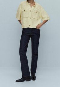 Żółty, teksturowany, dzianinowy top z krótkim rękawem, z dwiema przednimi kieszeniami i złotymi guzikami, w zestawieniu z ciemnoniebieskimi prostymi dżinsami i czarnymi botkami za kostkę.