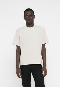 Ελαφρώς μπεζ κοντομάνικη μπλούζα με ριμπ υφή, στρογγυλή λαιμόκοψη και άνετη γραμμή, σε συνδυασμό με μαύρο παντελόνι.