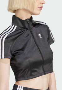Zwarte cropped sportjas met een hoge kraag, korte mouwen, witte strepen aan de zijkanten en een ritssluiting met een klein logo op de voorkant.