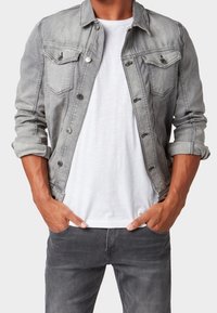Grå denimjacka med knapplås, fickor med klaff och uppvikta ärmar, bärs över en vit t-shirt och tillsammans med grå jeans.