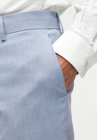 Χέρι στην τσέπη ανοιχτομπλε παντελονιού, φοριέται λευκό πουκάμισο με μακριά μανίκια και μανσέτα με κουμπιά, κοντινή λήψη στην περιοχή της μέσης και του ισχίου.